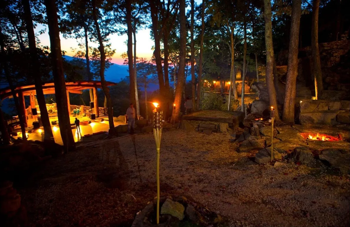 A cozy outdoor seating arrangement encircles a glowing fire pit, set among tall trees. The setup is ideal for evening conversations, storytelling, or simply enjoying the tranquility of nature. Torches add atmospheric lighting, enhancing the sense of warmth and togetherness.