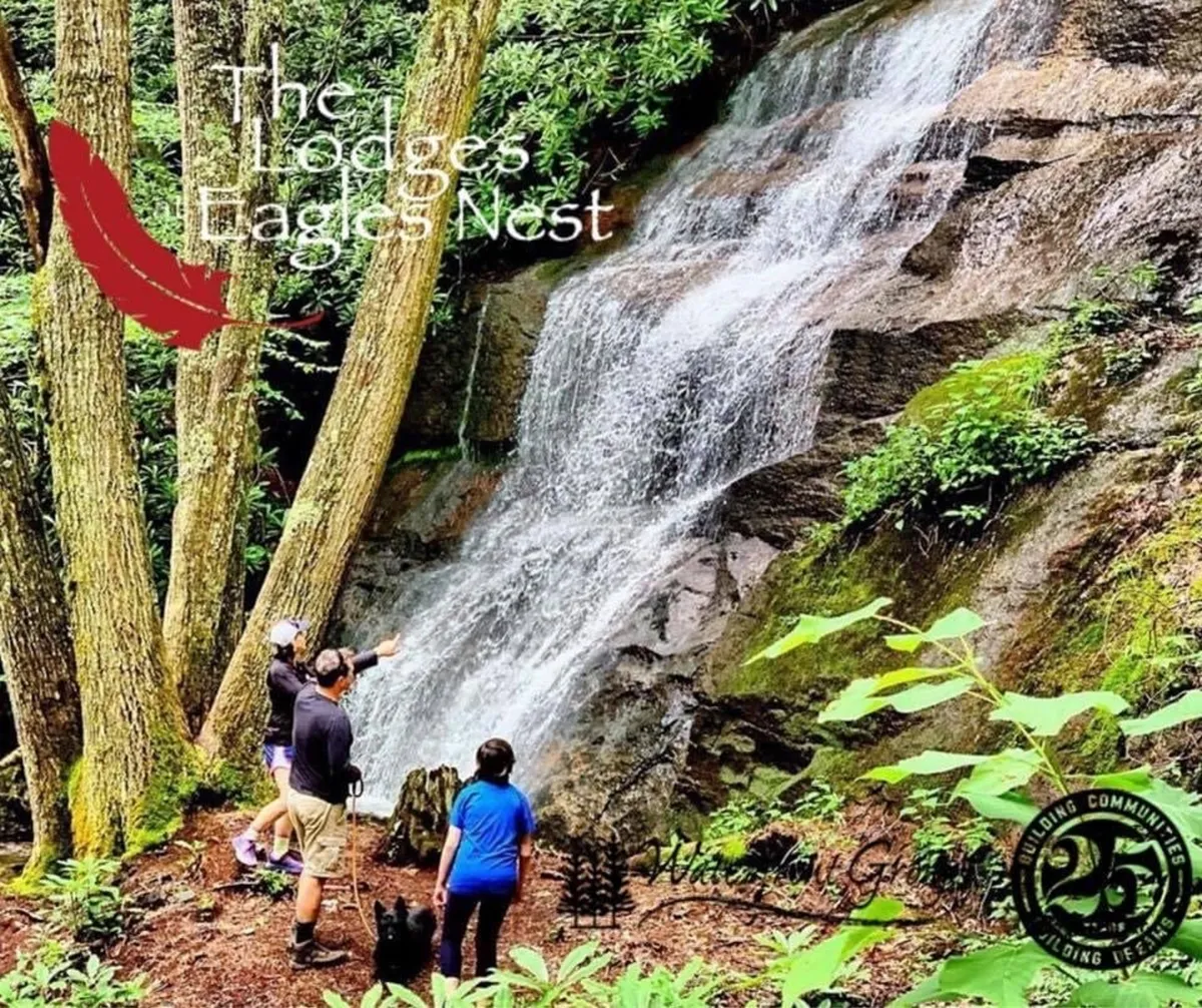 A family and their dog are seen hiking near a stunning waterfall, surrounded by lush forest. The group is exploring the natural landscape and enjoying the refreshing mist from the falls.