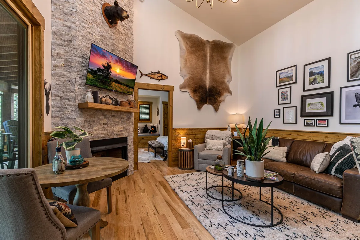 A living room featuring a prominent stone fireplace with a gas log fire, a mounted deer head above, a large flat screen TV, and comfortable seating arranged around a coffee table adorned with plants. The space exudes a rustic mountain ambiance, inviting guests to unwind for movie nights or cozy gatherings by the fire.