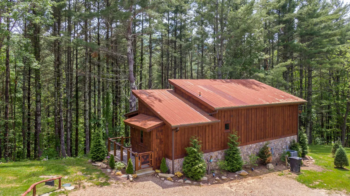 A peaceful exterior view of a wooden cabin with a stone foundation, surrounded by mature trees and set along a gravel driveway. The porch overlooks a natural setting, ideal for watching sunsets, enjoying wildlife sightings, and stargazing at night.