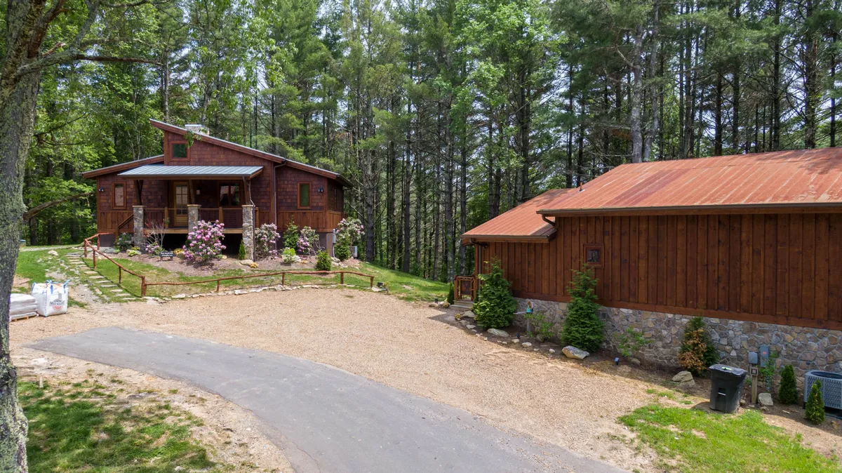 A rustic wooden cabin is surrounded by flowering plants and a gravel driveway, creating an atmosphere ripe for outdoor activities. The open porch area suggests comfortable spots for morning coffee, evening bonfires, or simply gathering as a group to enjoy nature together.