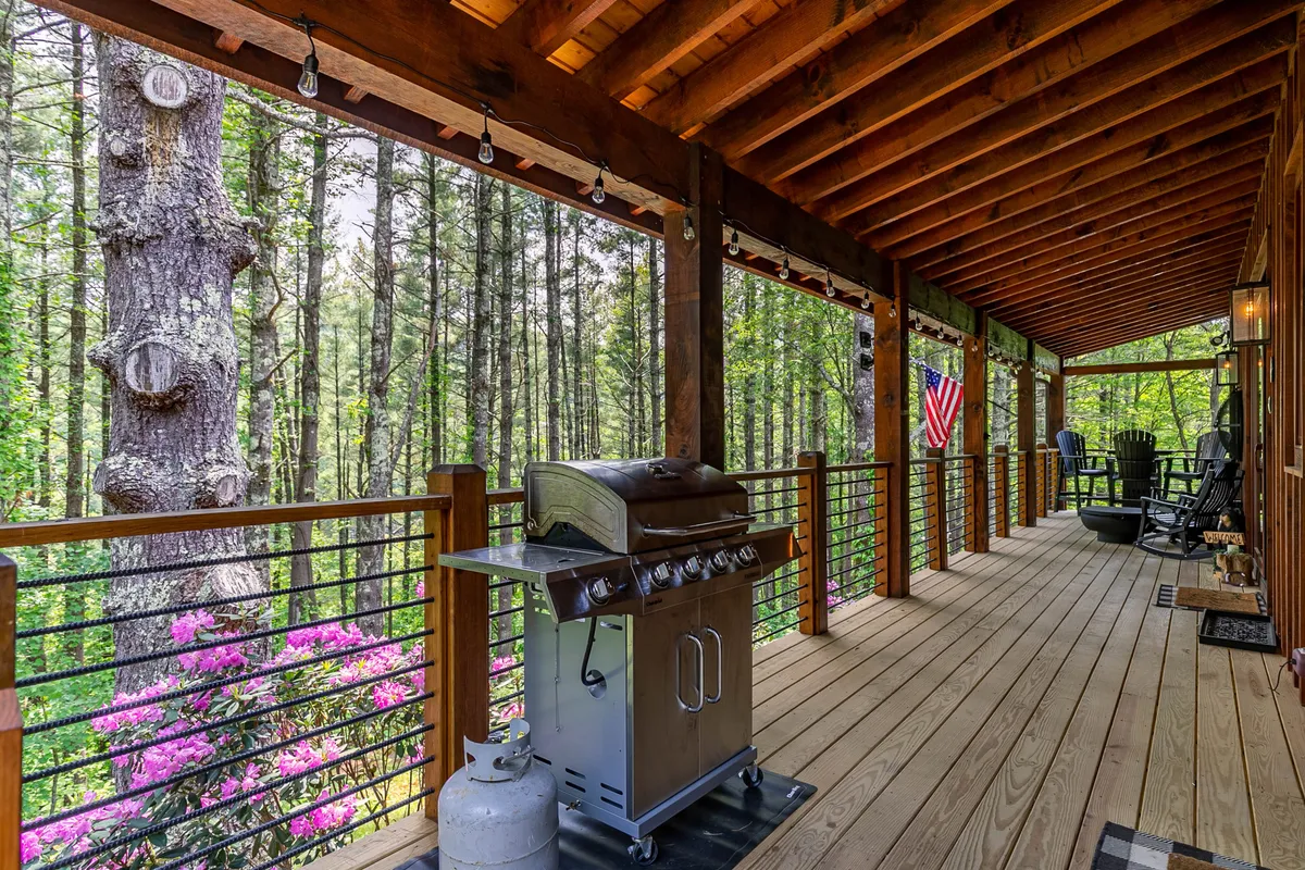 A welcoming outdoor grilling area with a grill, rocking chairs, American flag, and blooming flowers. The deck offers a cozy space for barbecues and gatherings, with sunset views over the trees.