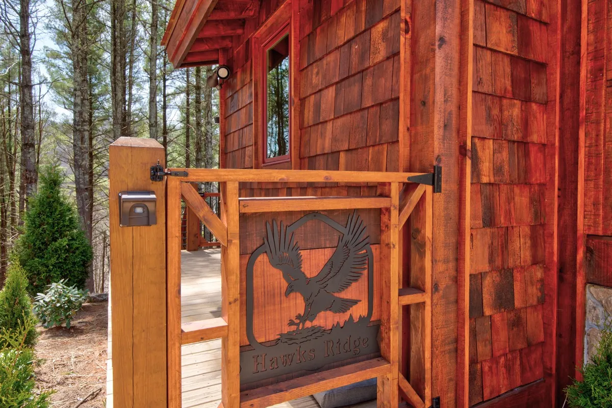 The image showcases a distinctive wooden gate with a hawk design, set against shingle siding and embraced by trees. The gate feels like an entryway into something special—hinting at both privacy and the sense of entering an exclusive, secure enclave.