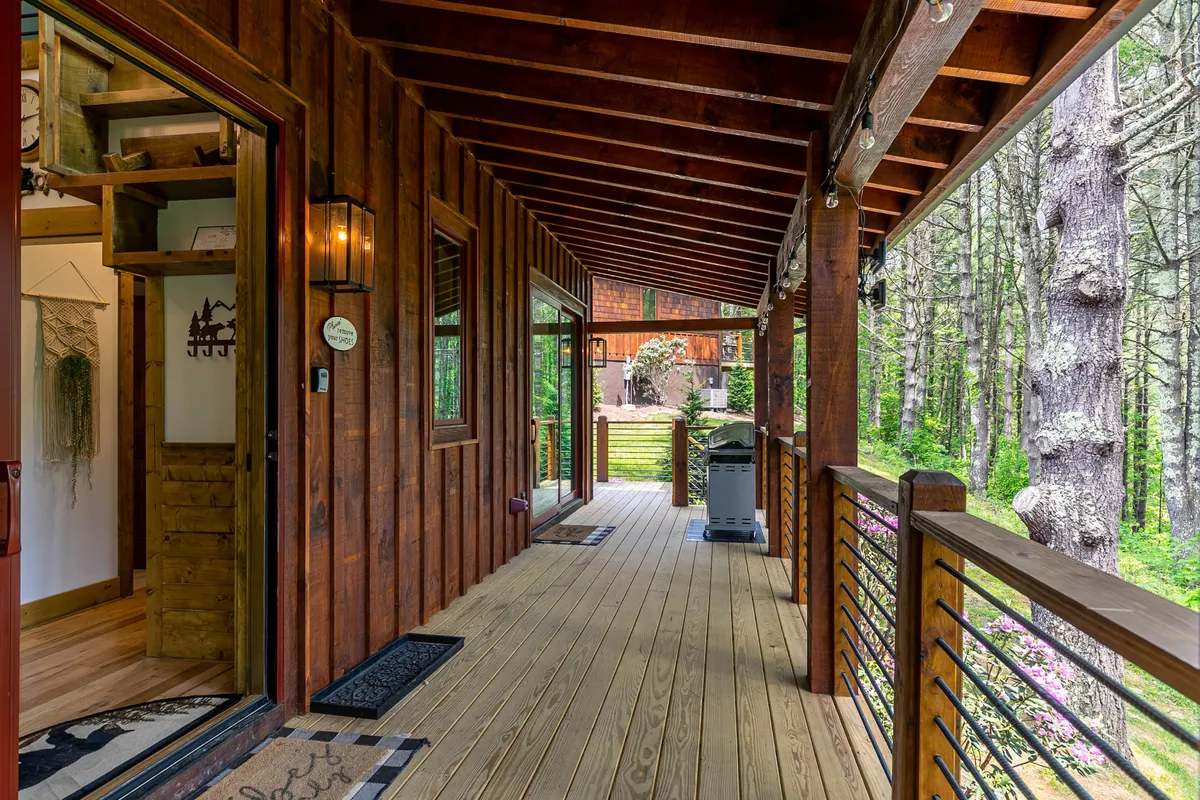 A welcoming deck scene featuring a wooden railing, outdoor grill, and comfortable seating, with the doorway leading directly back into the interior. The surrounding natural greenery envelops the space, suggesting a seamless transition between indoor and outdoor living. The deck is illuminated softly, hinting at the potential for both morning relaxation and evening gatherings under string lights.