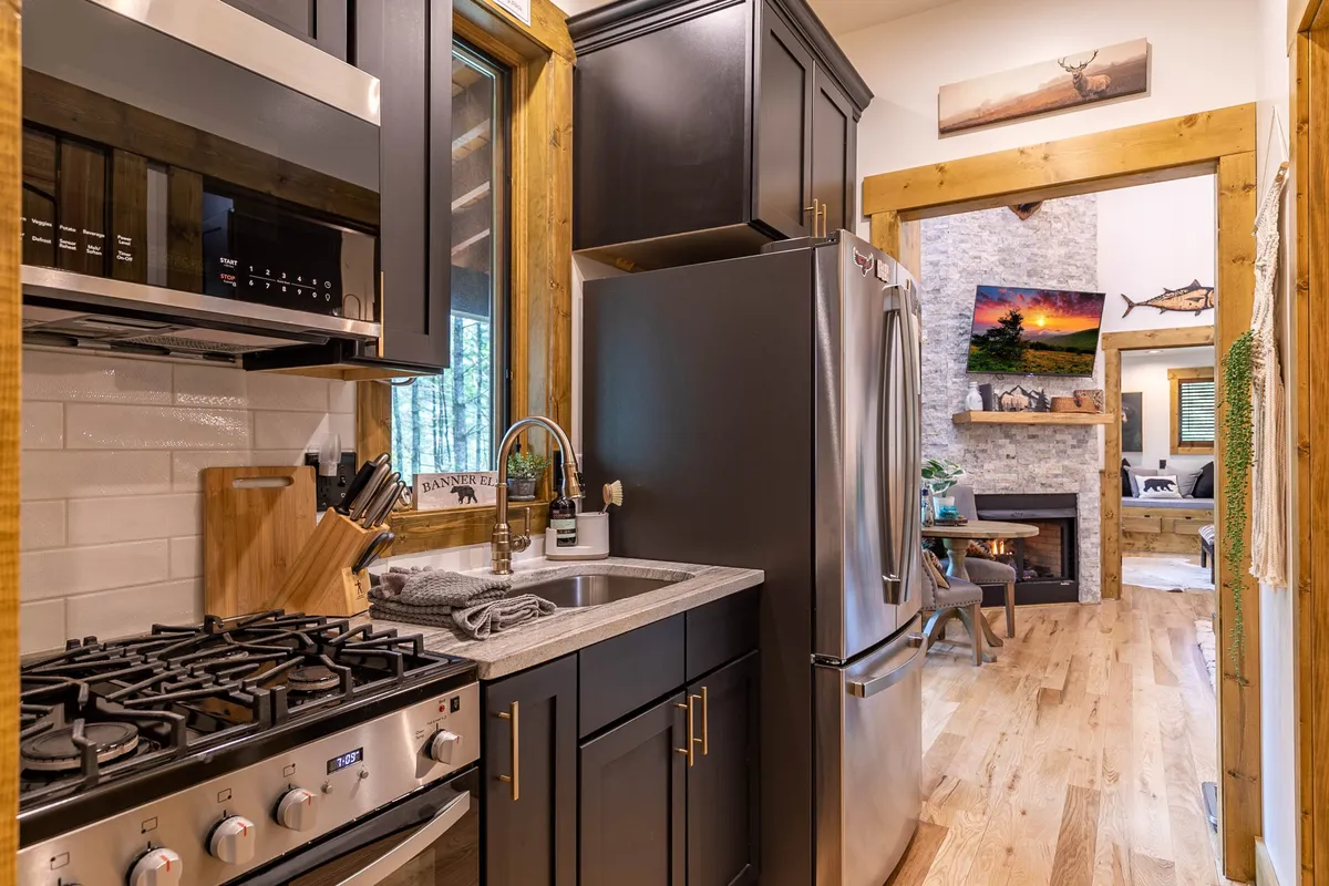 A modern kitchen with stainless steel appliances, a granite countertop, dark cabinetry, and a large window letting in natural light. The scene captures the essence of a well-equipped kitchen ready for cooking feasts or sharing morning coffee, as well as the open flow to the adjacent living area.
