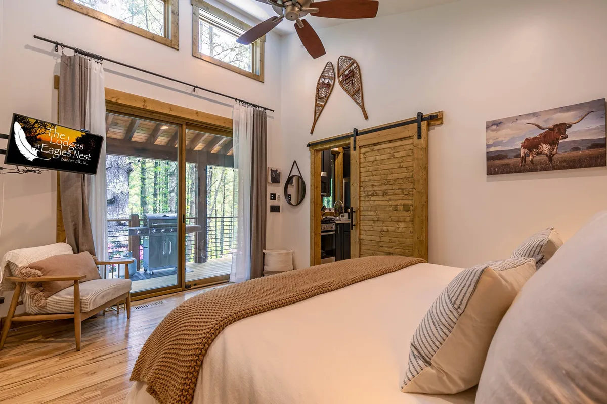 This image showcases a serene bedroom with neutral bedding, large glass doors leading to a balcony, a rustic wooden barn door, decorative snowshoes on the wall, and wall art of a longhorn. Visible features include a television and a chair for relaxing or reading, while the glass doors suggest direct deck access and a tranquil connection to the outdoors.