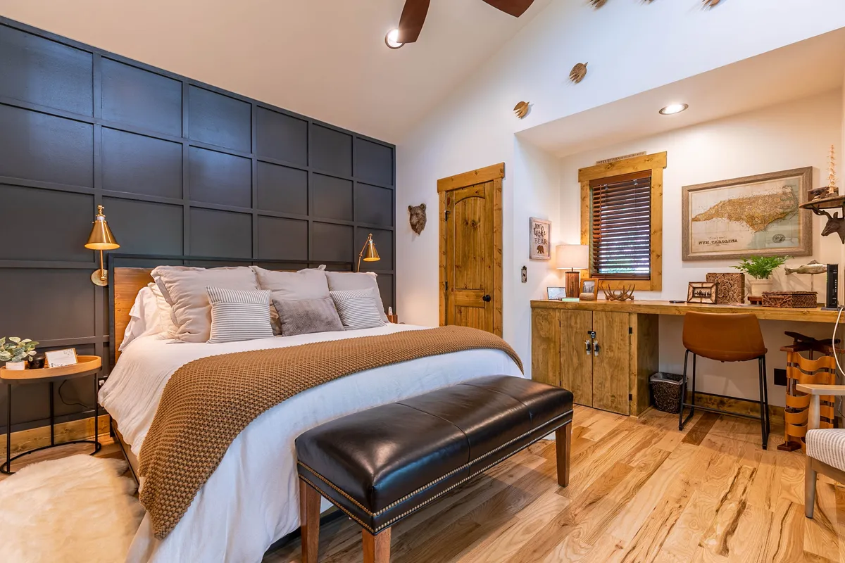 A warmly decorated bedroom featuring a bed with decorative pillows, a wooden desk, an accent wall, and sunlight filtering through window blinds. The cozy setup suggests a perfect spot for relaxation, reading, or working, with the desk positioned for views outside.