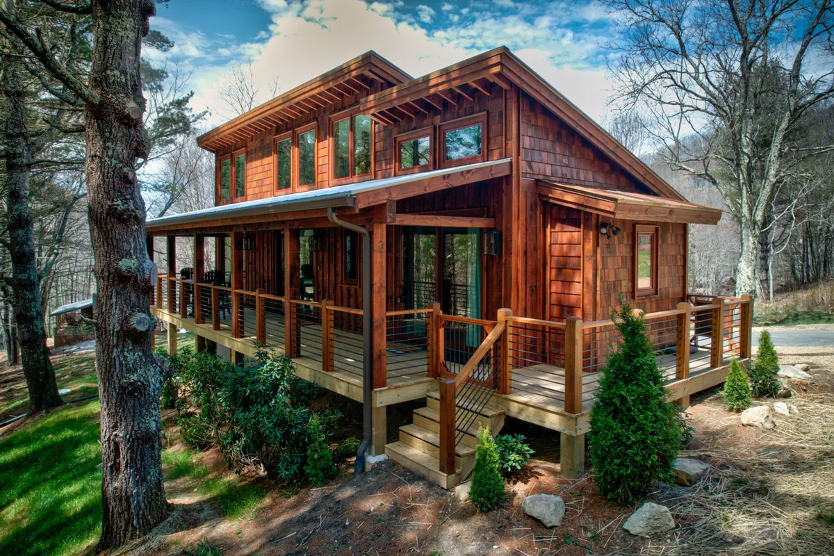The image showcases the warm wooden exterior of the cabin, Hawks Ridge, nestled among towering trees in the gated community of Fish Camp II. Large windows offer glimpses of the inviting interior, and a spacious front porch sets the scene for relaxing with morning coffee or watching evening sunsets over the forested landscape.
