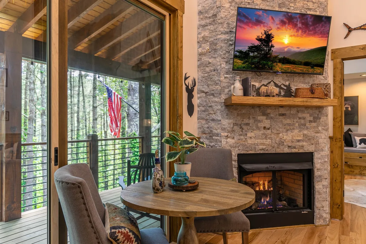 A wide shot of an open living and dining area featuring a fireplace, TV, dining table, and large windows offering views of the outdoors. This perspective reveals how the living, dining, and kitchen spaces are interconnected, underscoring the open-plan design that encourages communal activities and easy conversation.