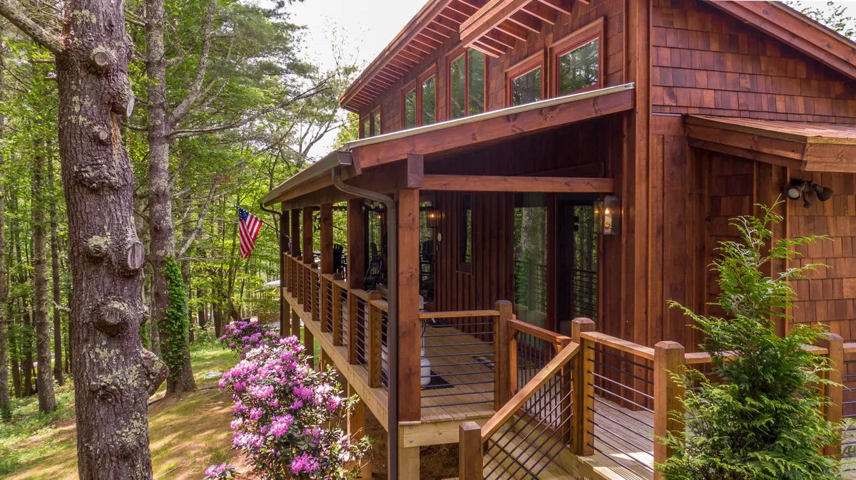 This image features a welcoming wooden deck adorned with flowers, large windows that provide a glimpse into the indoors, and an American flag waving gently in the breeze. The setting suggests a space perfect for socializing or relaxing outdoors, surrounded by serene nature.