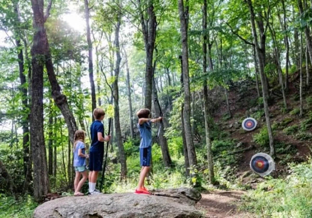 This image shows children practicing archery outdoors, aiming at target boards set against a picturesque rock formation, surrounded by trees. The kids are actively engaged, and the scene captures an energetic, family-friendly outdoor activity.